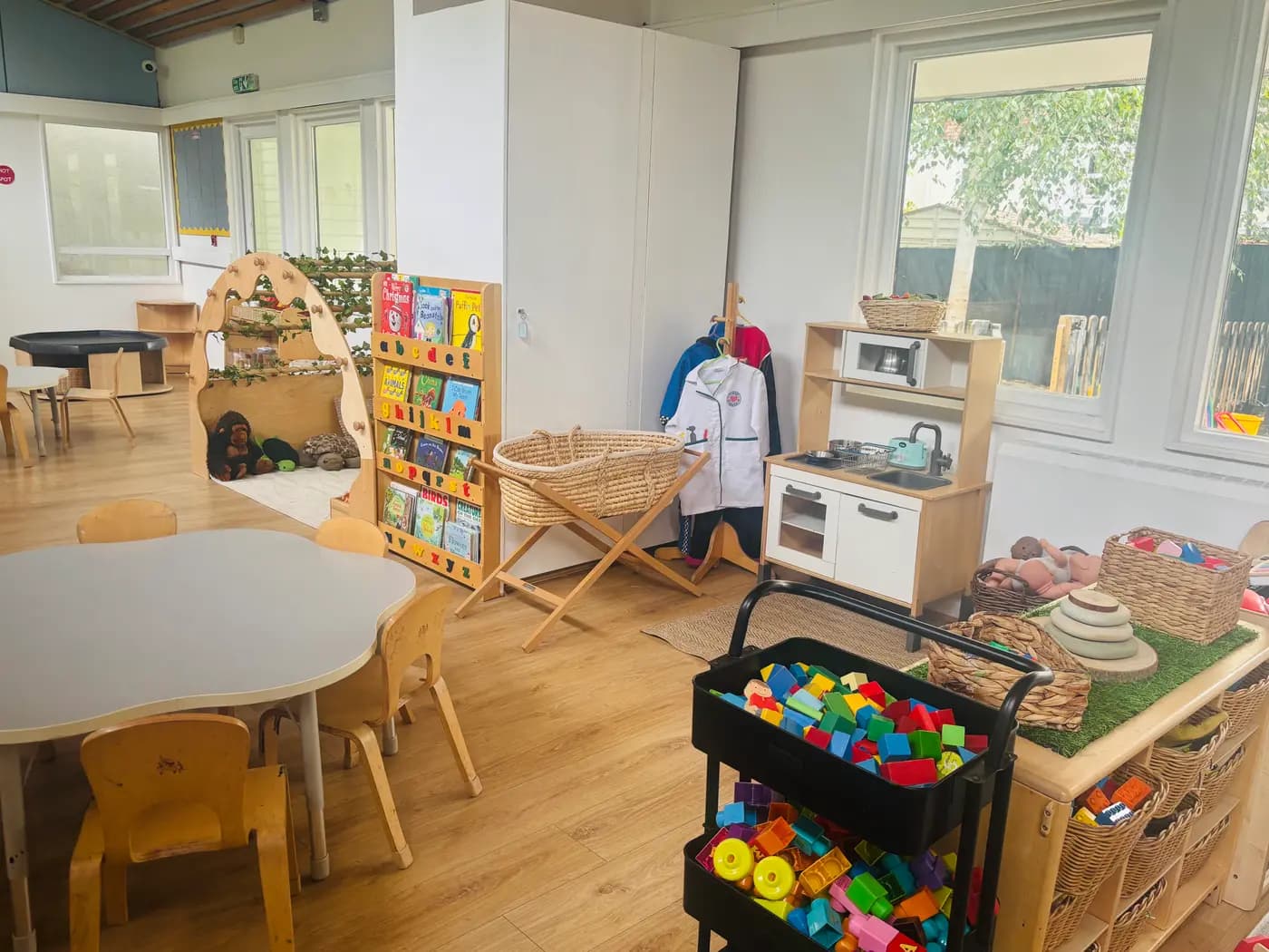 The preschool room with a book display, moses basket, dress-up rail and play kitchen at Blue Nest Montessori Aldershot