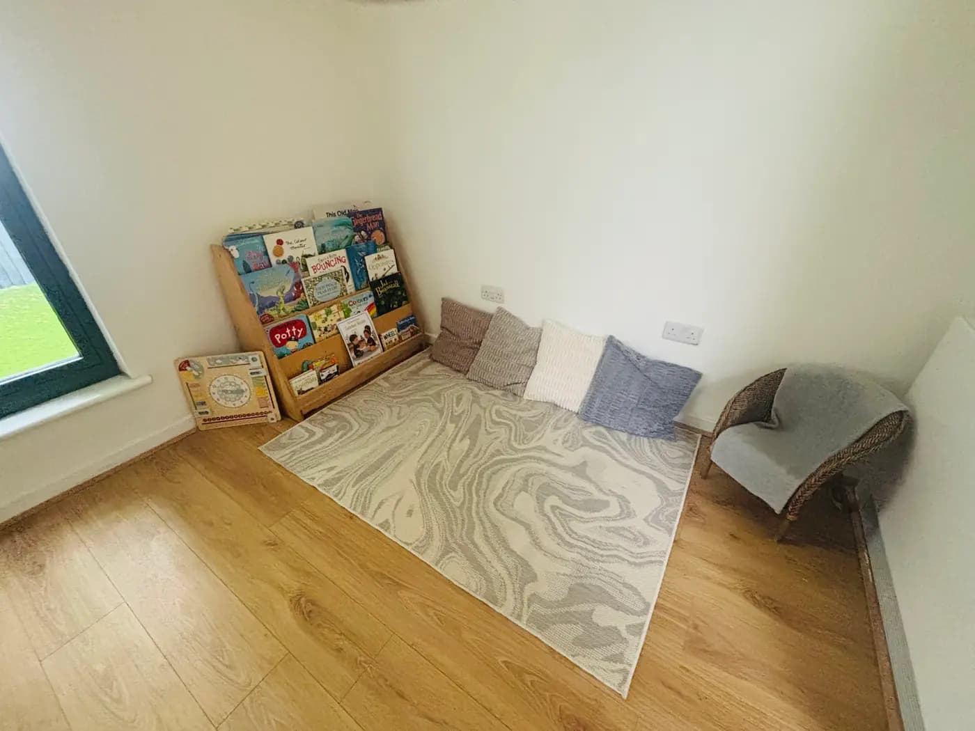 A quiet reading corner with cushions, a bookshelf and a wicker chair at Blue Nest Montessori Aldershot